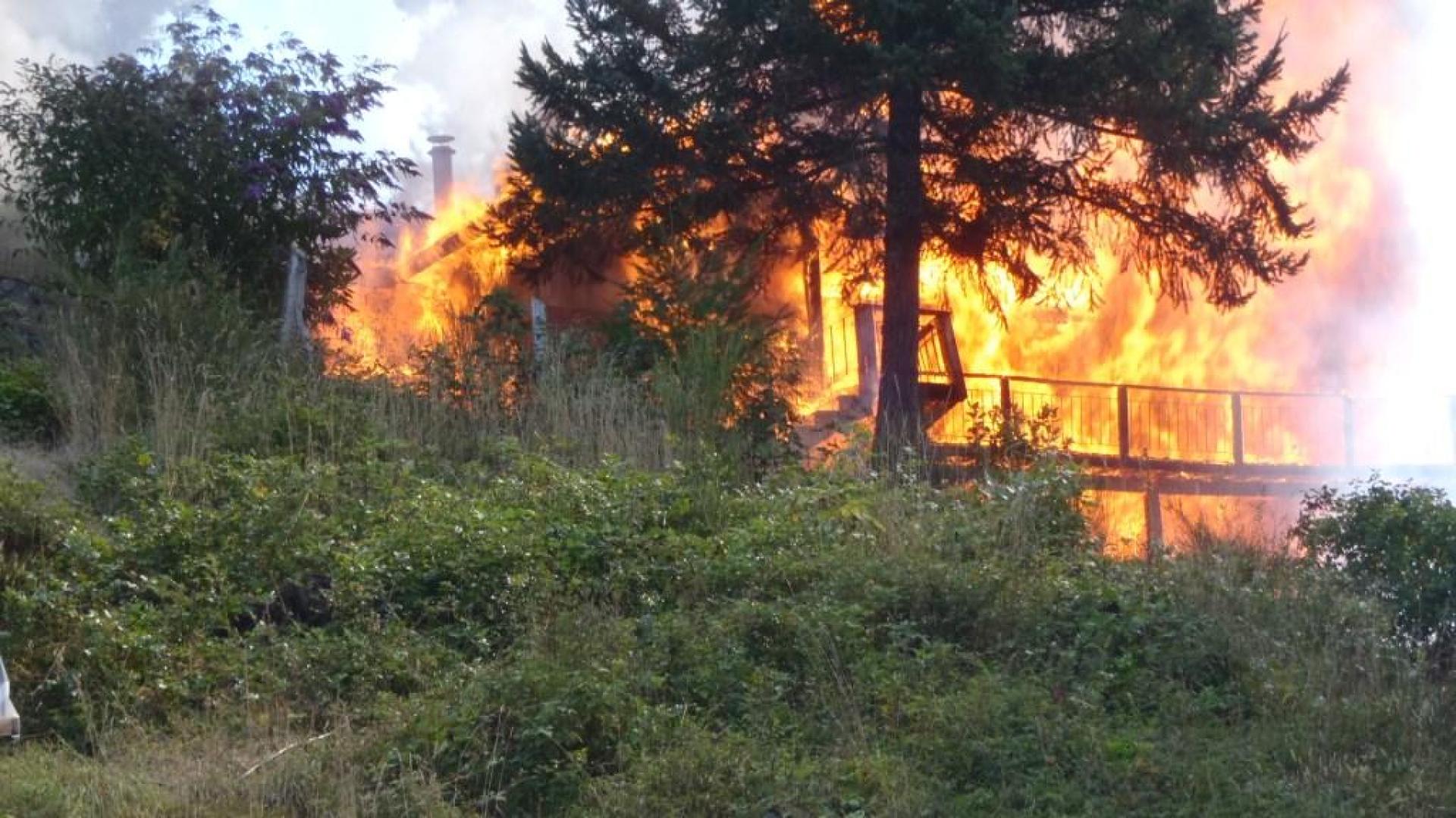 Wooden structure on fire. The fire is surrounded by greenery.